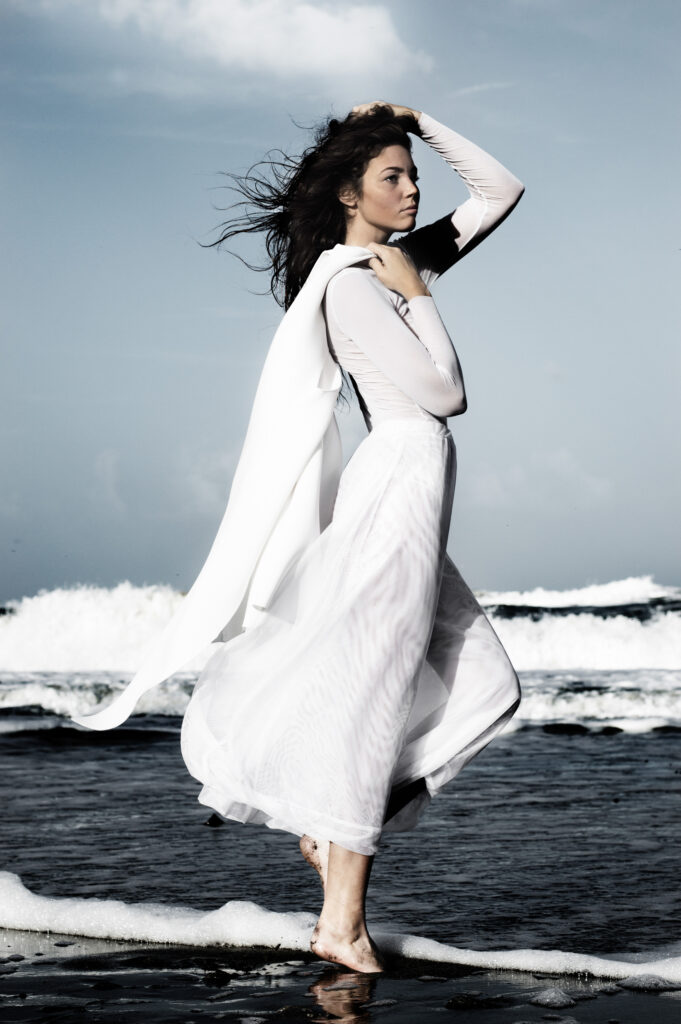 Woman standing on the beach with the sun illuminating her white dress and wind blowing her white dress.